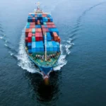 Top-down view of a cargo ship at sea, with colorful shipping containers stacked on deck and a visible wake behind it.